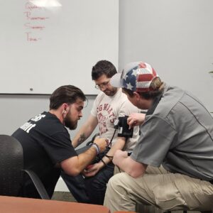 An EMT instructor inside an ambulance demonstrates proper technique on an infant manikin for a group of young children, providing a public outreach learning experience.