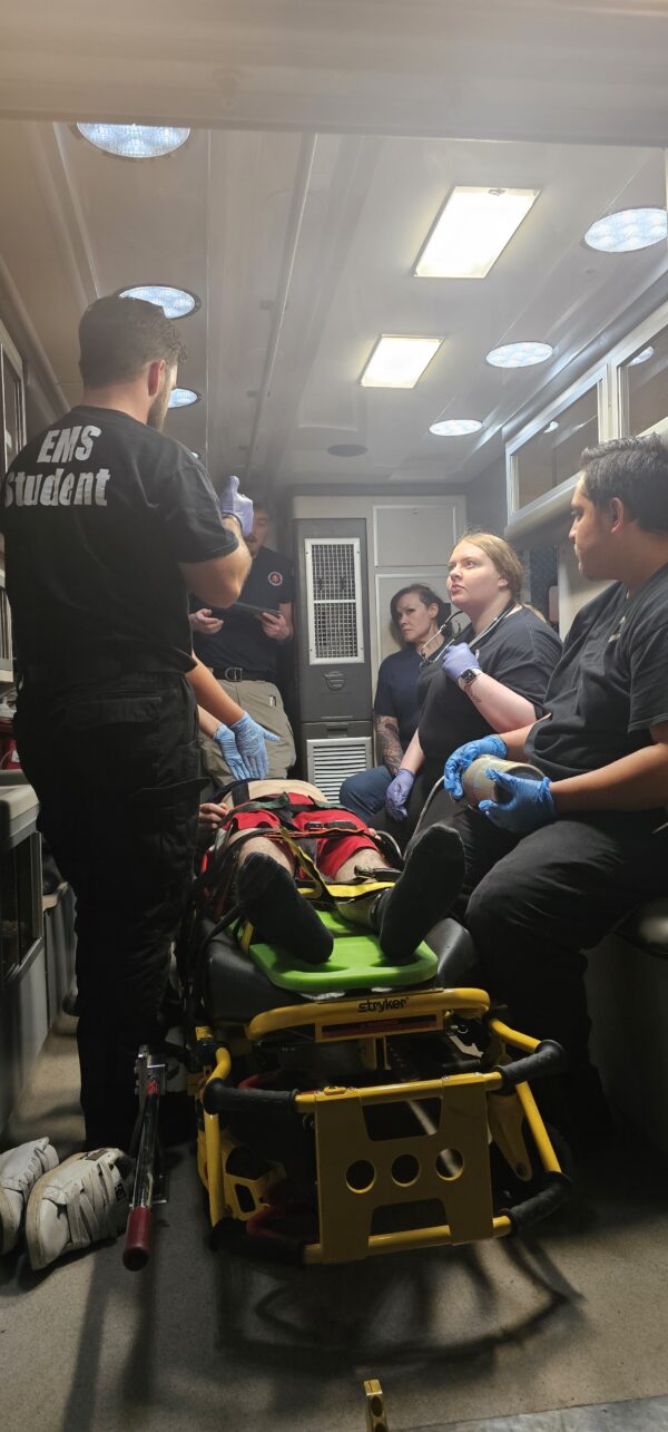 Three male EMT students in a classroom practice taking a manual blood pressure reading on one another as part of their hands-on training.