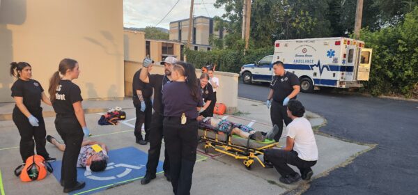 EMT students conduct a training simulation of a car accident, with one student playing a patient in a cervical collar while others prepare a backboard for a safe extrication.